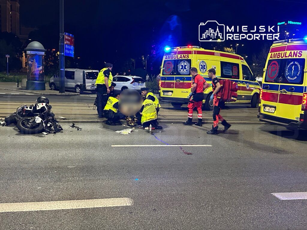 Motocykl leży na jezdni, a ratownicy medyczni udzielają pomocy poszkodowanemu w pobliżu ambulansów i policji.