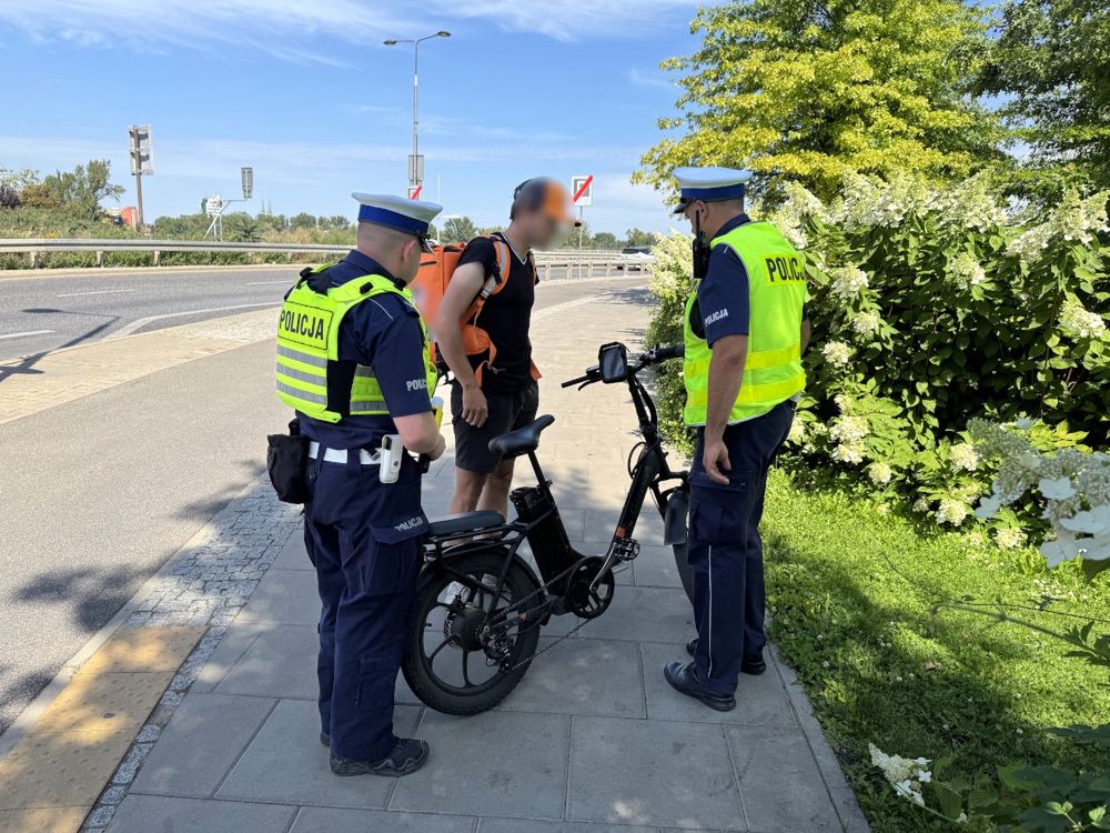 Dwóch policjantów w kamizelkach odblaskowych rozmawia z mężczyzną przy rowerze elektrycznym na chodniku.