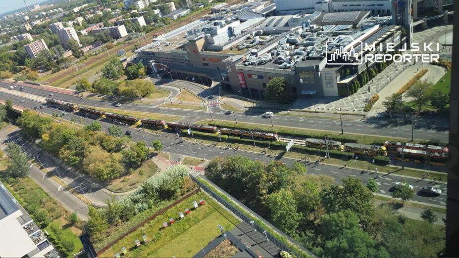 Widok z góry na ruchliwą ulicę z tramwajami, obok nowoczesnego centrum handlowego otoczonego zielenią.