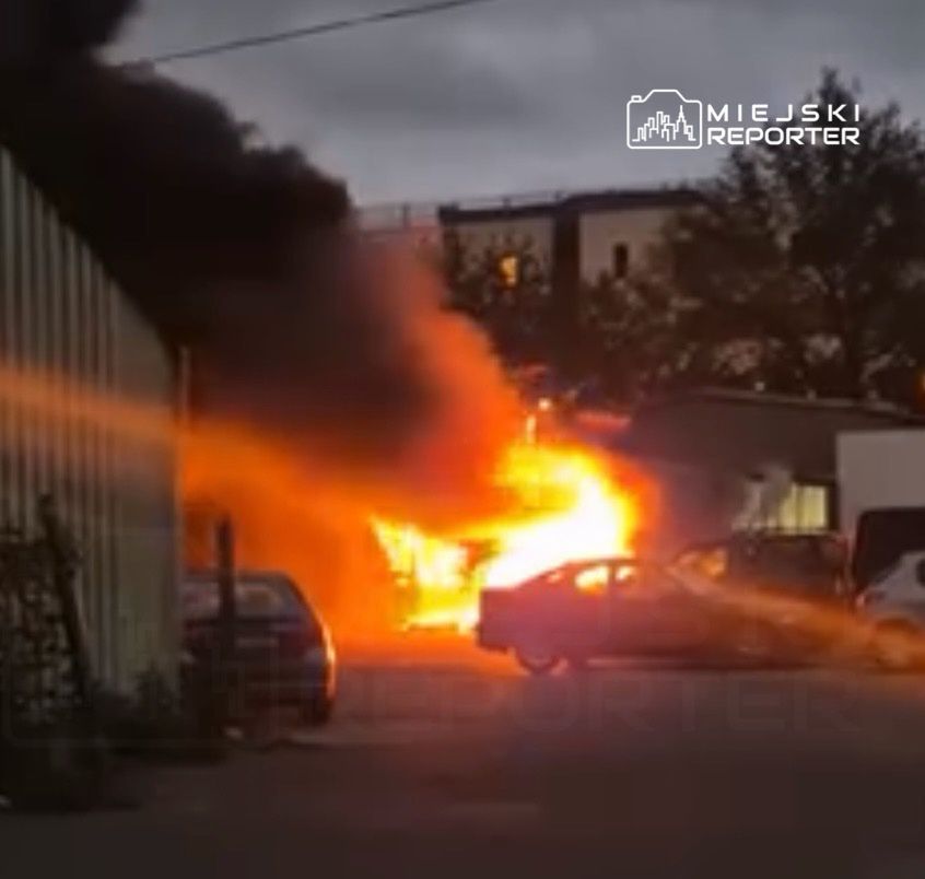 Pojazd w płomieniach na parkingu, z dużą ilością czarnego dymu unoszącego się w powietrze.