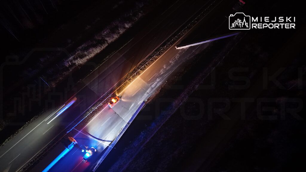 Policyjny radiowóz z włączonymi sygnałami świetlnymi stoi na drodze, obok leżącego przewróconego słupa.