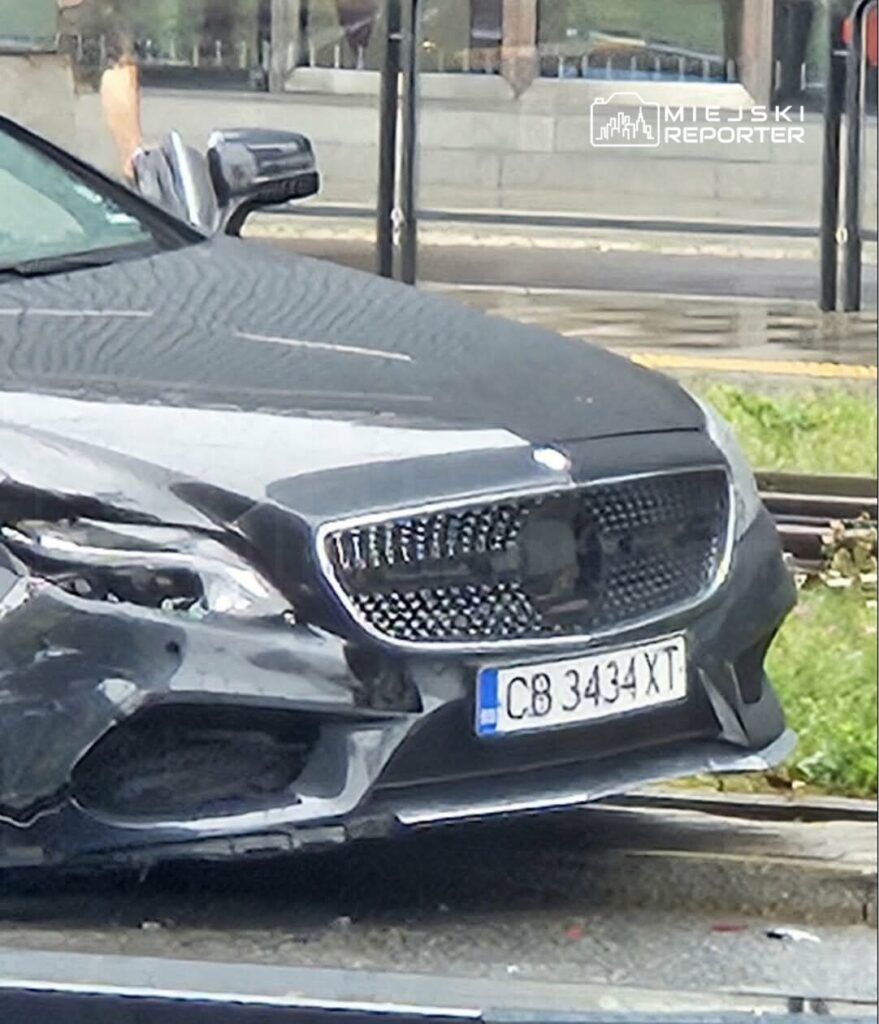 Czarny samochód osobowy z uszkodzeniami przód stoi na ulicy obok przystanku komunikacyjnego.