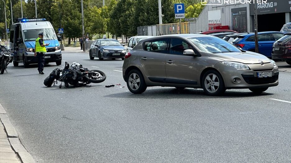 Motocykl leży na jezdni obok srebrnego samochodu, a policjant sprawdza sytuację w pobliżu karetki.