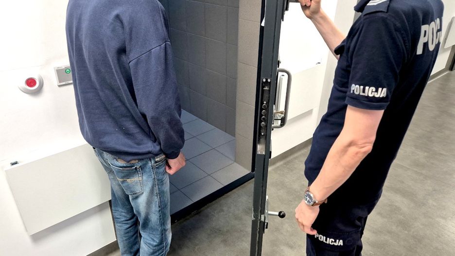 Mężczyzna w dresie stoi w pomieszczeniu, a policjant otwiera drzwi prowadzące do celi. W tle widać szare płytki.