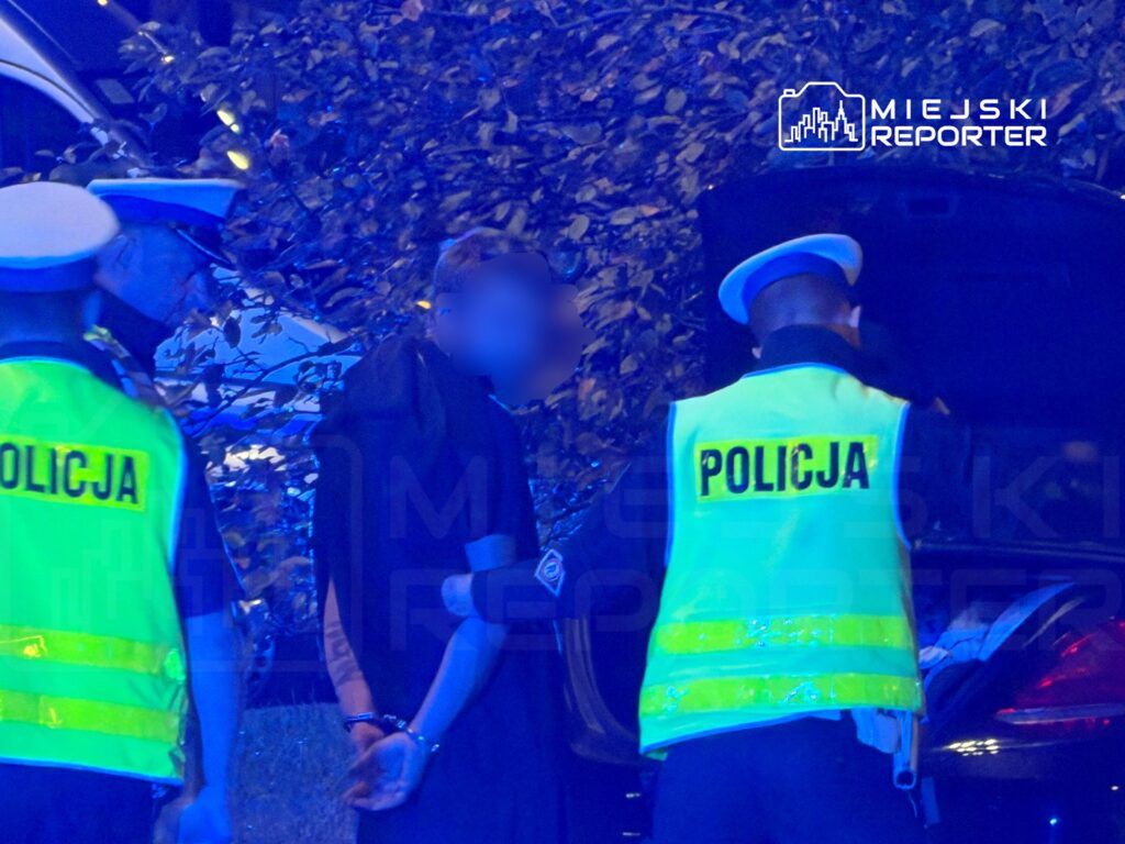 Mężczyzna w czarnej odzieży z rękami skrępowanymi w kajdankach stoi obok samochodu, otoczonego przez policjantów w kamizelkach.