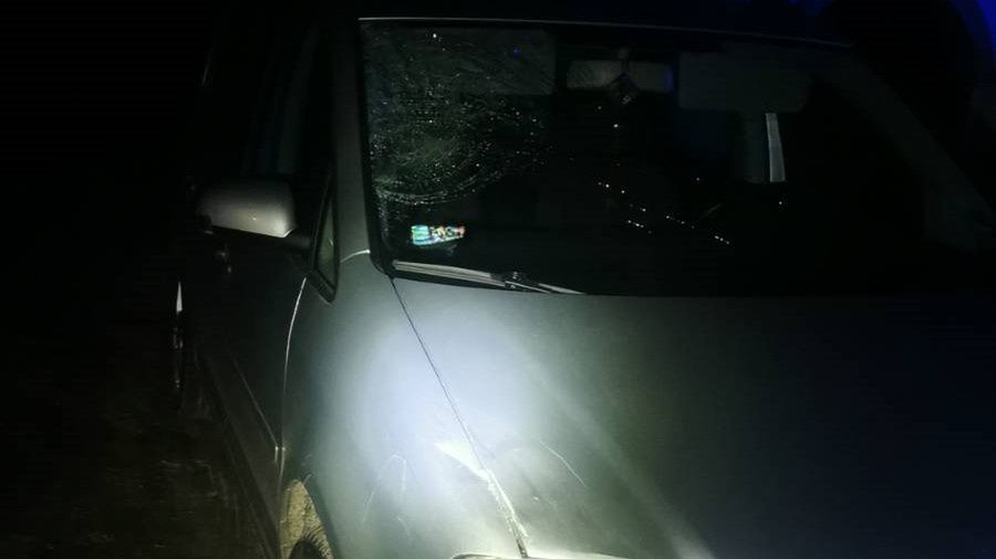 Uszkodzony samochód osobowy z pękniętą szybą przednią, zaparkowany w ciemnym otoczeniu z niebieskim oświetleniem.