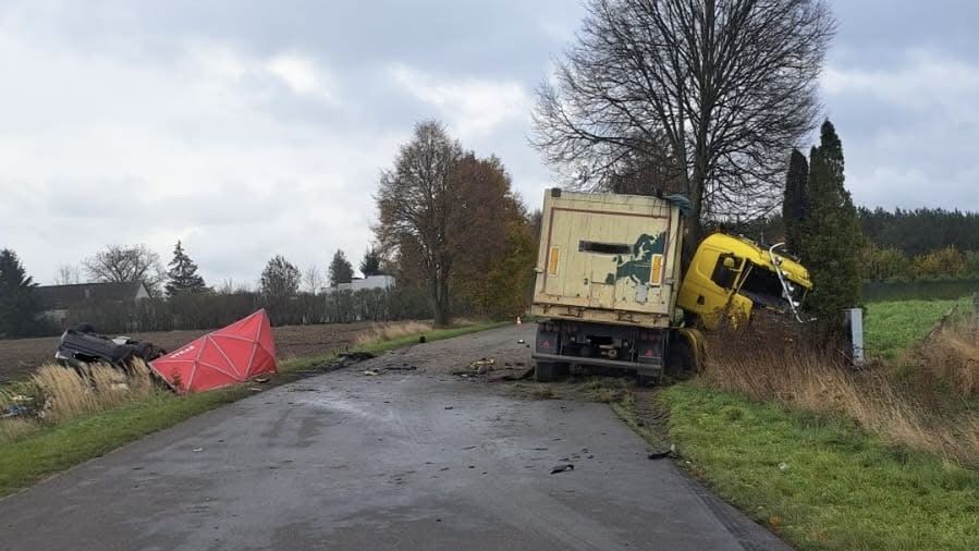 Złomowany żółty ciężarówka w rowie, obok przewrócony czerwony namiot na wiejskiej drodze otoczonej drzewami.
