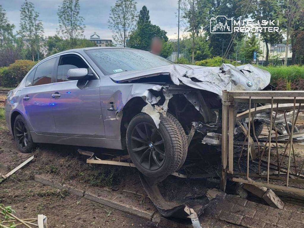 Uszkodzony samochód osobowy marki BMW wjeżdża w ogrodzenie, leżąc na stoku w otoczeniu zieleni.