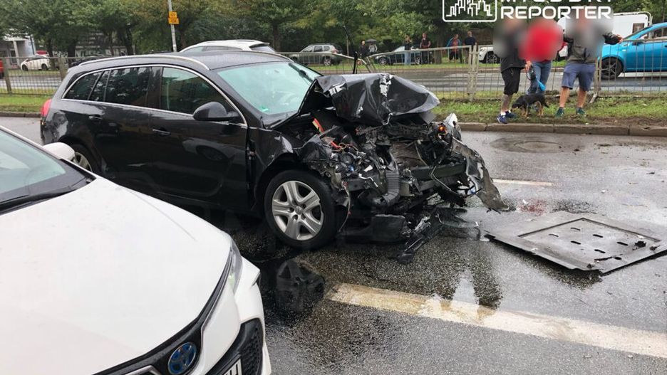 Zniszczony samochód osobowy z uszkodzoną przednią częścią stoi na mokrej jezdni, obok grupy ludzi obserwujących zdarzenie.