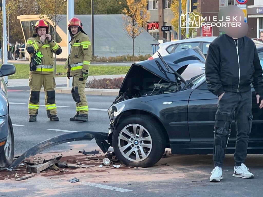 Dwóch strażaków w hełmach obserwuje uszkodzony samochód osobowy z otwartą maską na ruchliwej ulicy.
