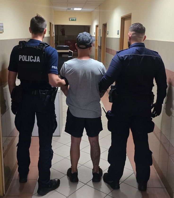 Dwaj policjanci prowadzą mężczyznę w szarym T-shircie i krótkich spodniach korytarzem w budynku.
