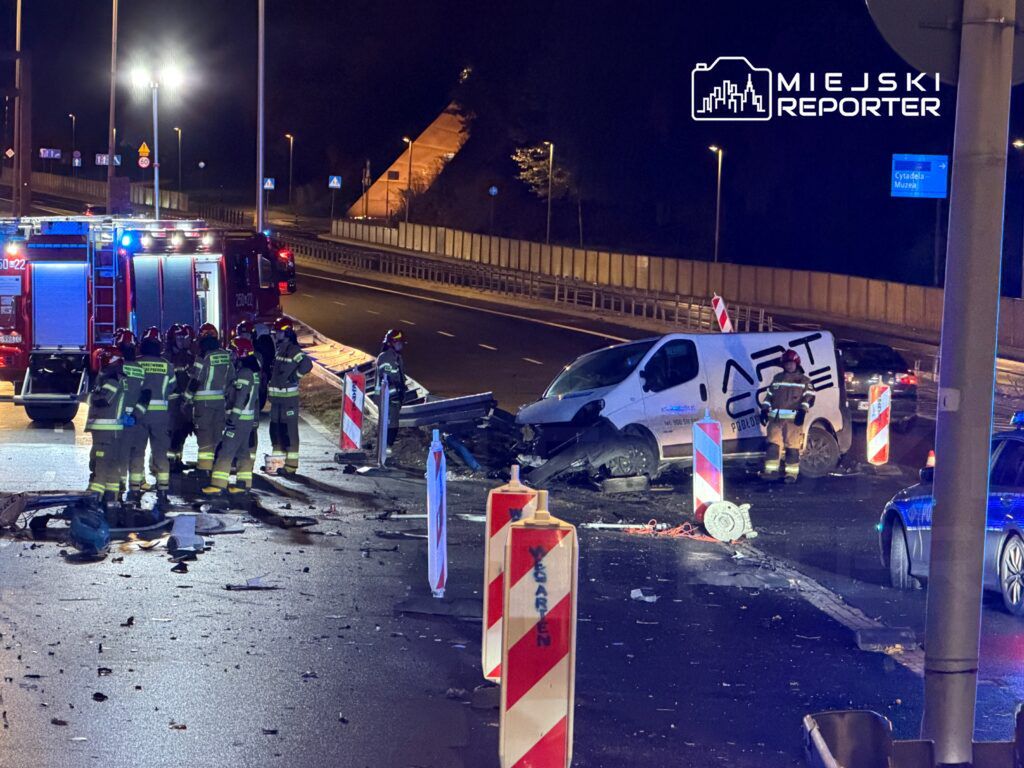 Grupa strażaków w mundurach bada miejsce wypadku drogowego, obok uszkodzonego furgonu na oświetlonej ulicy.