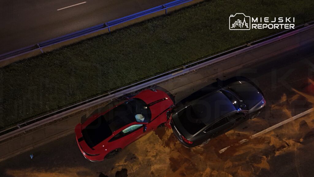 Czerwony samochód osobowy zderzony z ciemnym pojazdem na drodze, w tle widoczna trawa i barierka.