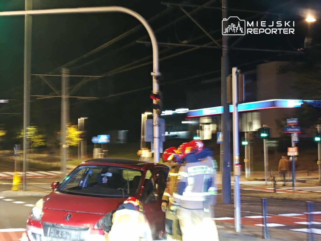 Czerwony samochód zatrzymany na skrzyżowaniu, obok trzech strażaków w mundurach, podczas nocnej interwencji.
