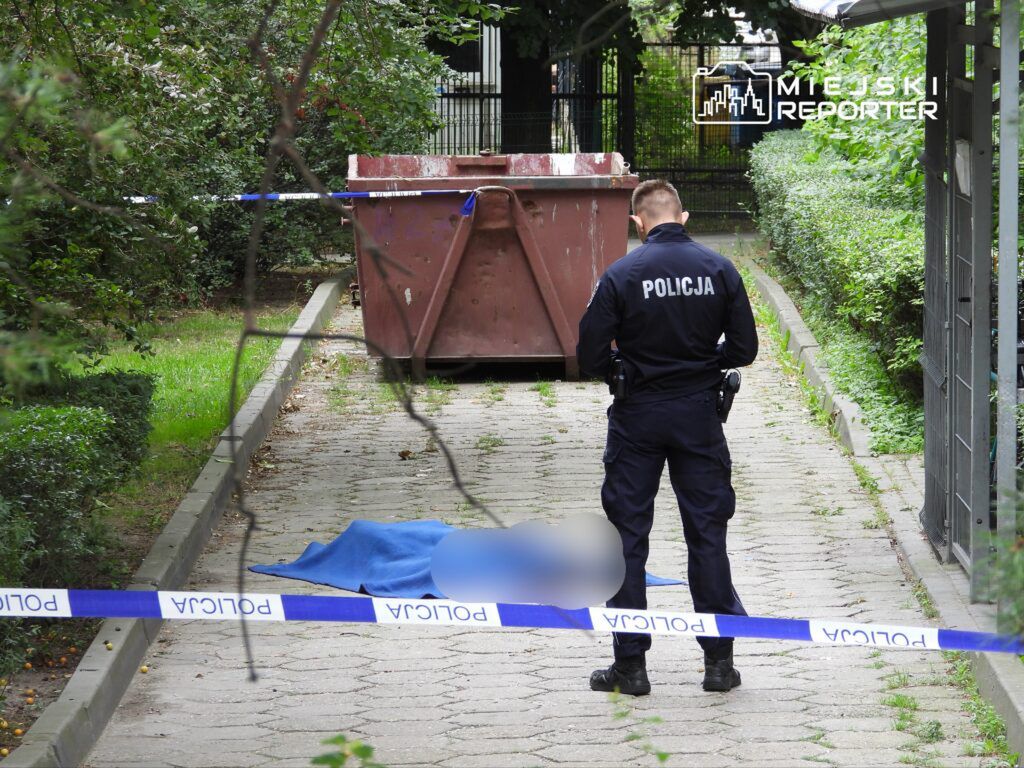 Policjant w mundurze bada miejsce zdarzenia przy kontenerze na śmieci, zabezpieczonym taśmą policyjną.