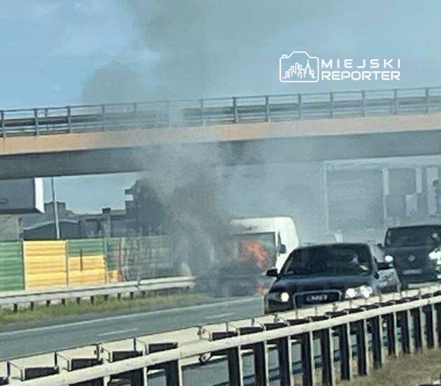 Dym i ogień wydobywają się z tyłu białego dostawczego samochodu na autostradzie, obok ruchu innych pojazdów.