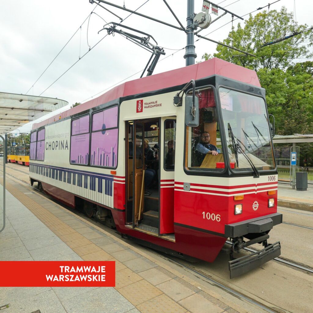 Tramwaj w barwach czerwono-białych zatrzymany na przystanku, z pasażerami wsiadającymi i wysiadającymi.