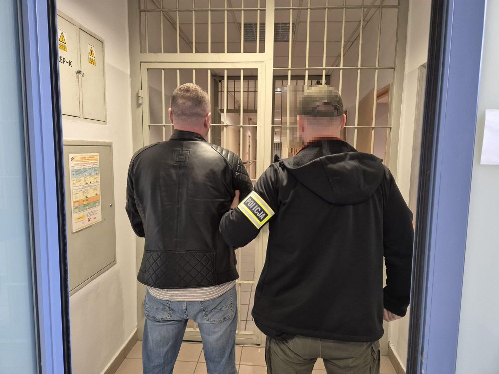 Mężczyzna w skórzanej kurtce prowadzony przez funkcjonariusza policji w kierunku zamkniętych drzwi aresztu.