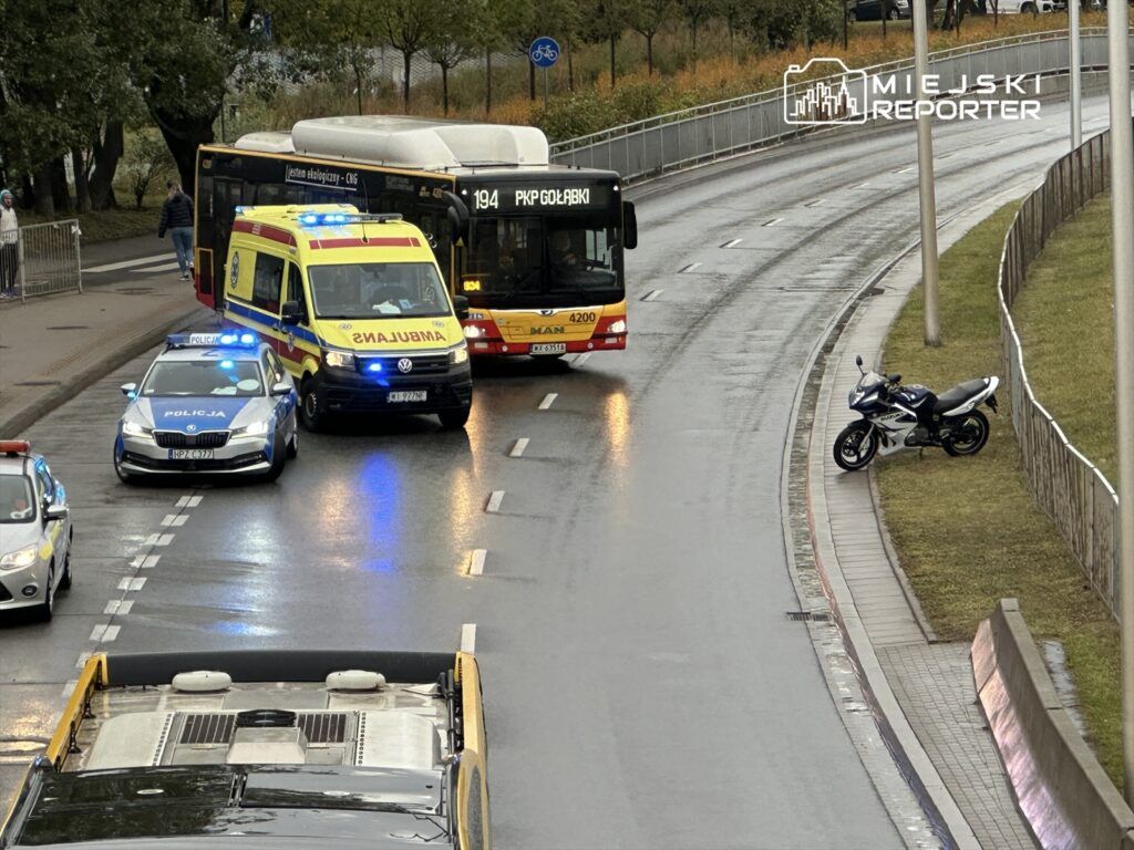 Na skrzyżowaniu zatrzymane są pojazdy: karetka z włączonymi sygnałami, radiowóz oraz autobus linii 194. Obok stoi motocykl.