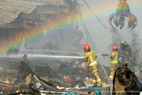 Dwaj strażacy w hełmach gaszą pożar zniszczonego budynku, w tle widać tęczę nad dymem.