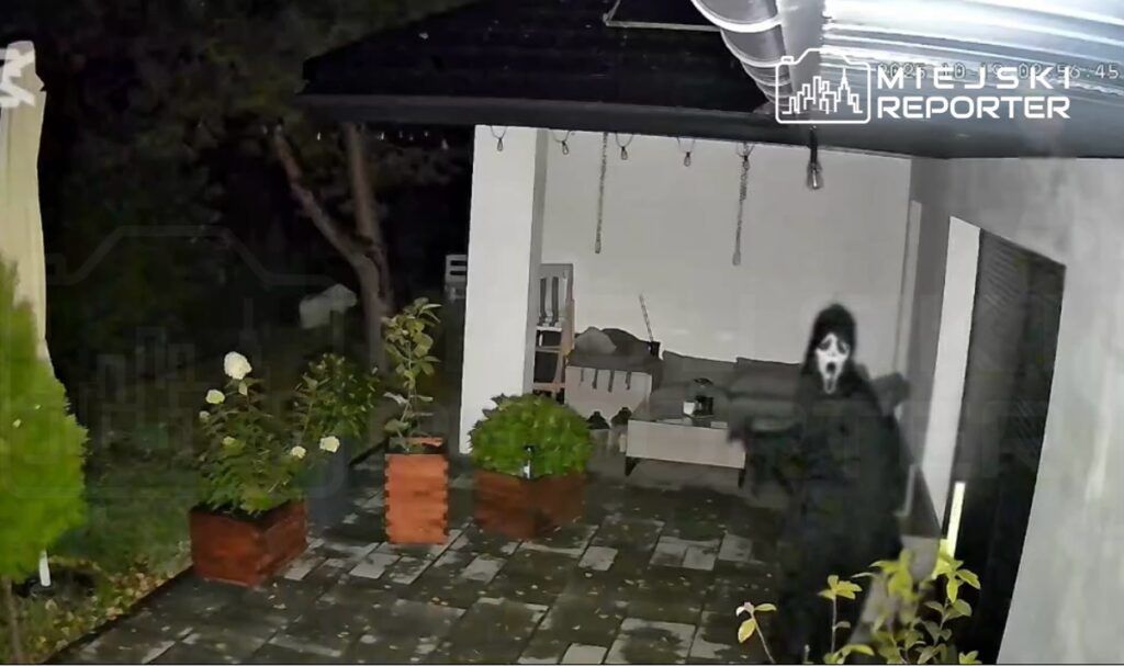 Osoba w czarnej pelerynie z maską przebrana za ducha stoi na patio, w tle widoczne rośliny i meble ogrodowe.