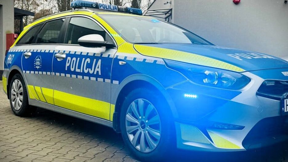 Radiowóz policyjny z niebieskimi i żółtymi oznaczeniami zaparkowany na kostce brukowej przed budynkiem.