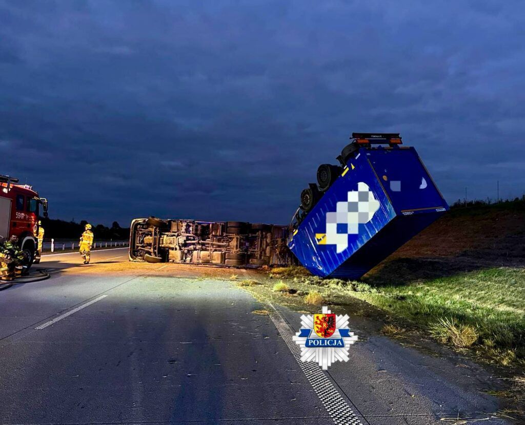 Na drodze leży przewrócony niebieski kontener, obok stoją strażacy i woz strażacki w tle, w ciemnej scenerii.