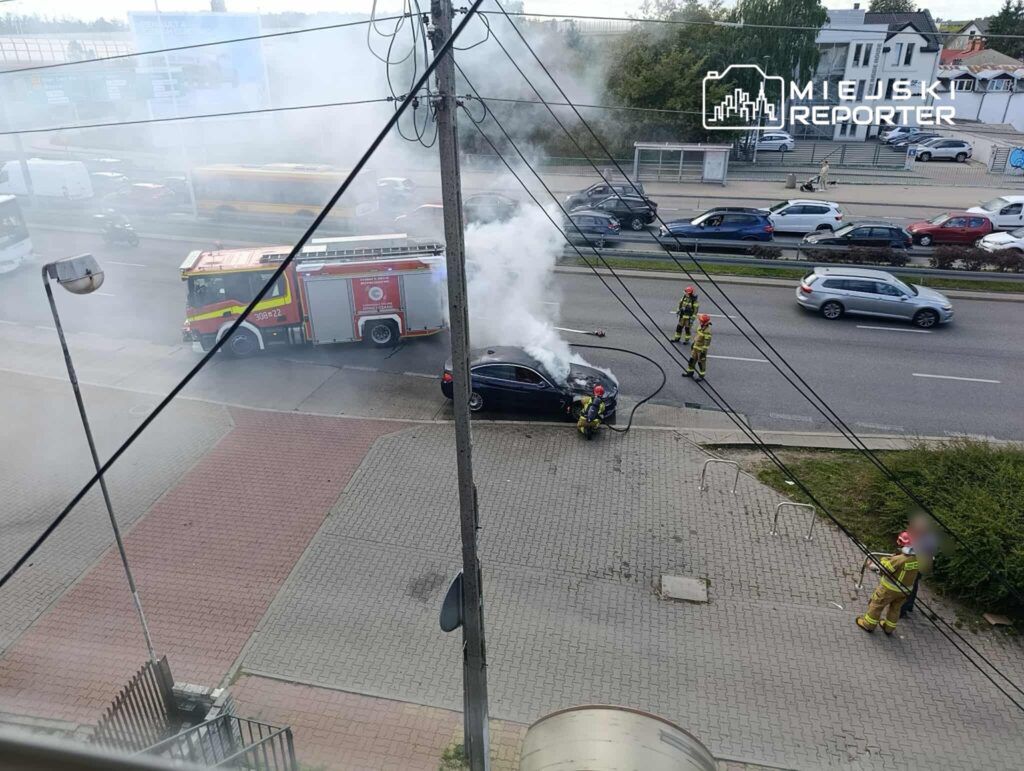 Strażacy gaszą pożar samochodu na chodniku obok ruchliwej ulicy, obok przejeżdżających pojazdów.