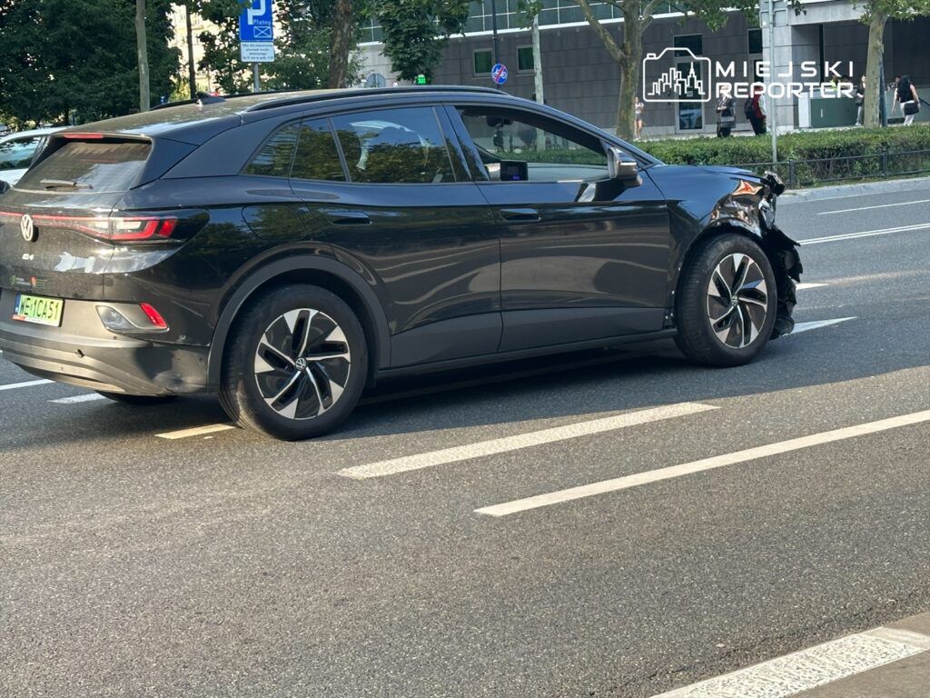 Czarny samochód osobowy z uszkodzoną przednią częścią stoi na jezdni w miejskim otoczeniu.