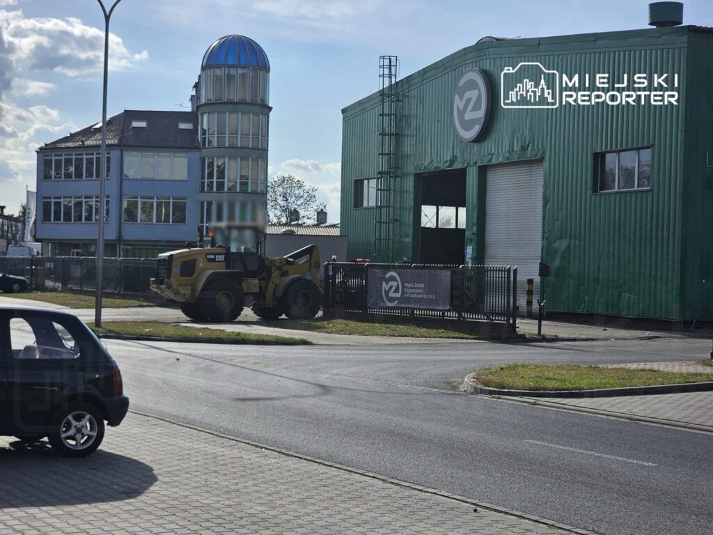 Żółty sprzęt budowlany podnosi materiały przed zielonym budynkiem przemysłowym z logo na fasadzie.