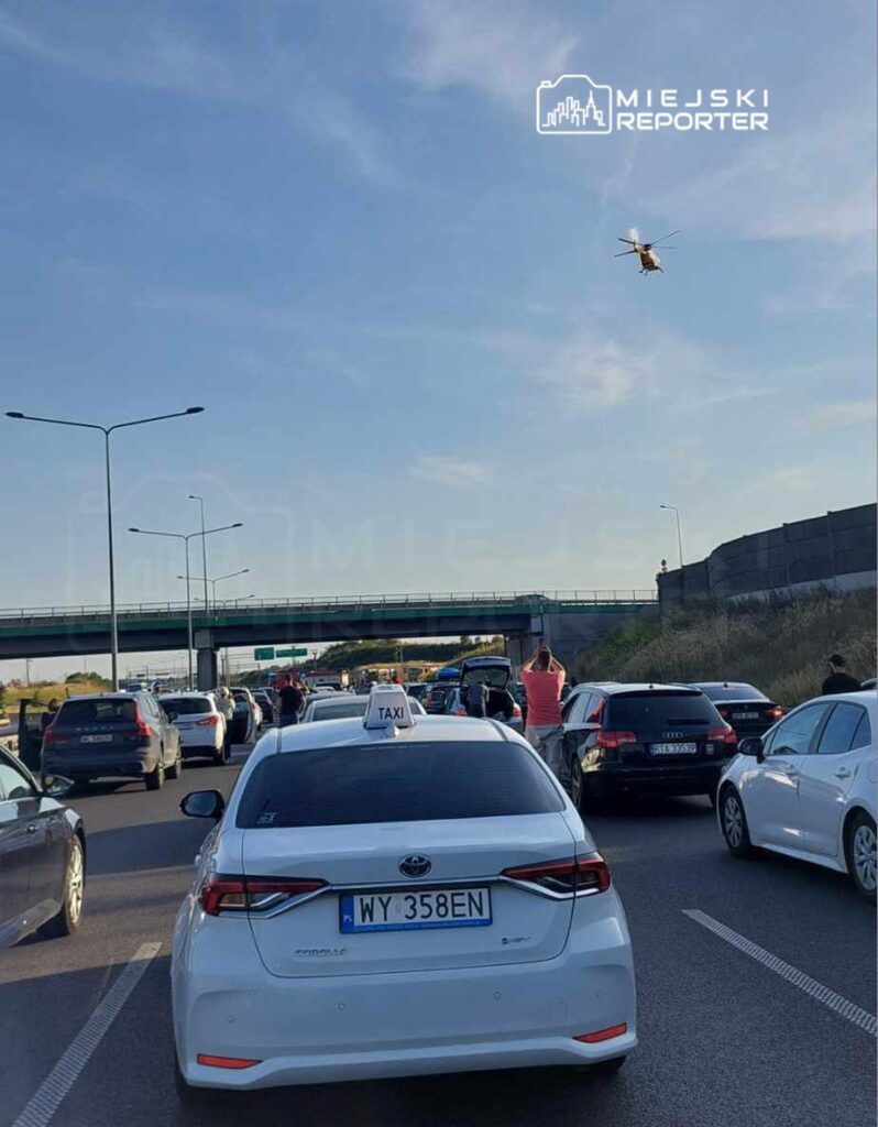 Ruch samochodowy zatrzymany na drodze, z białym taksówką na pierwszym planie i helikopterem unoszącym się w tle.