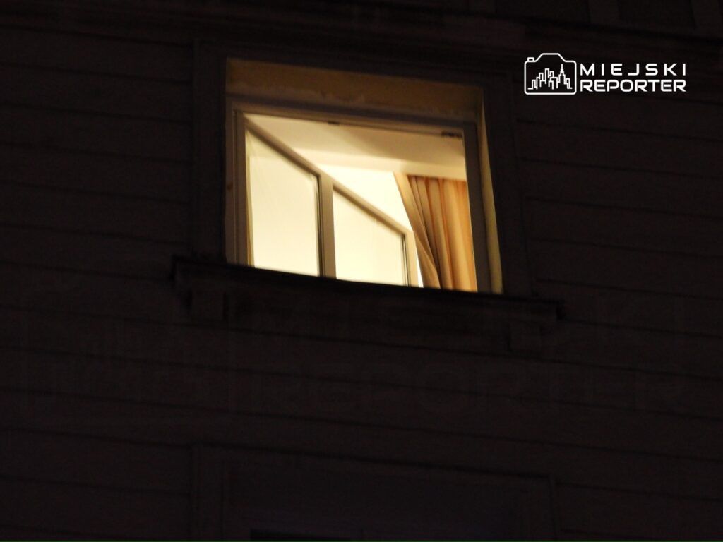 Oświetlone okno z zasłonami, otwarte na zewnątrz, w ciemnej elewacji budynku.