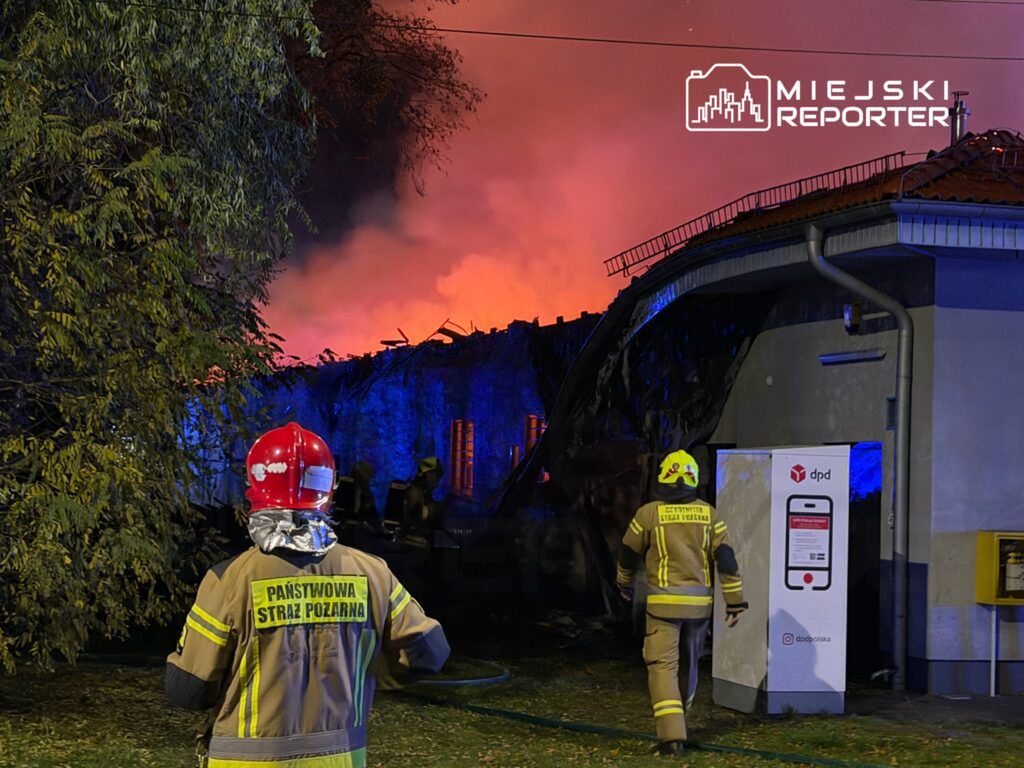 Strażacy w hełmach gaszą płonący budynek, w tle widoczne ogniste niebo i dym.