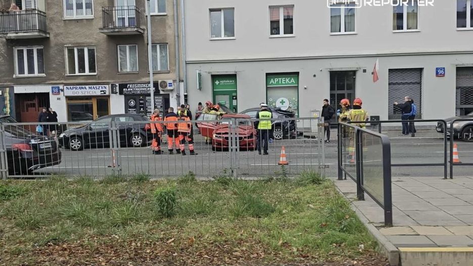 Grupa ratowników i policjantów pracuje na miejscu wypadku drogowego przy ulicy, obok zaparkowanych samochodów.