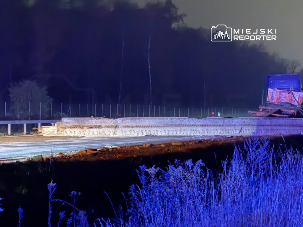 Droga budowlana z betonowymi elementami w tle, oświetlona niebieskim światłem, w otoczeniu niskiej roślinności.