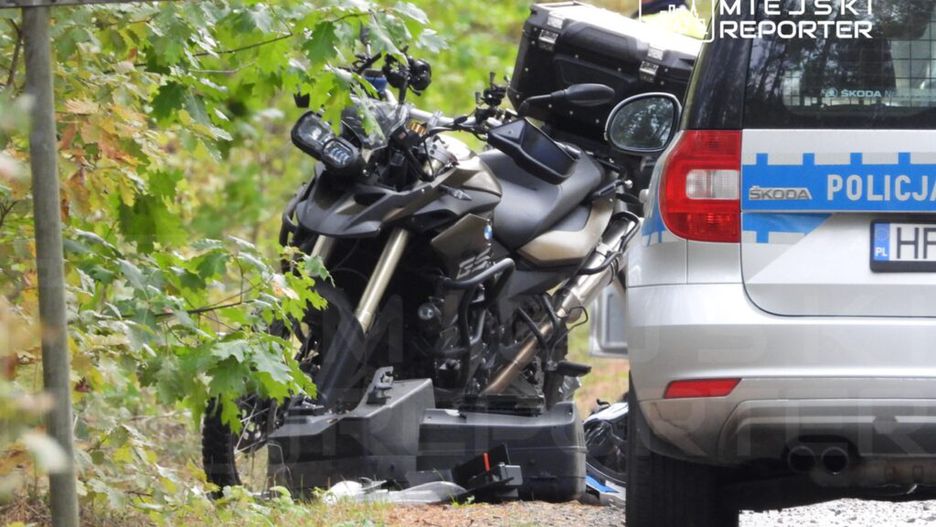 Motocykl leży na poboczu drogi w lesie, obok stoi radiowóz policyjny.