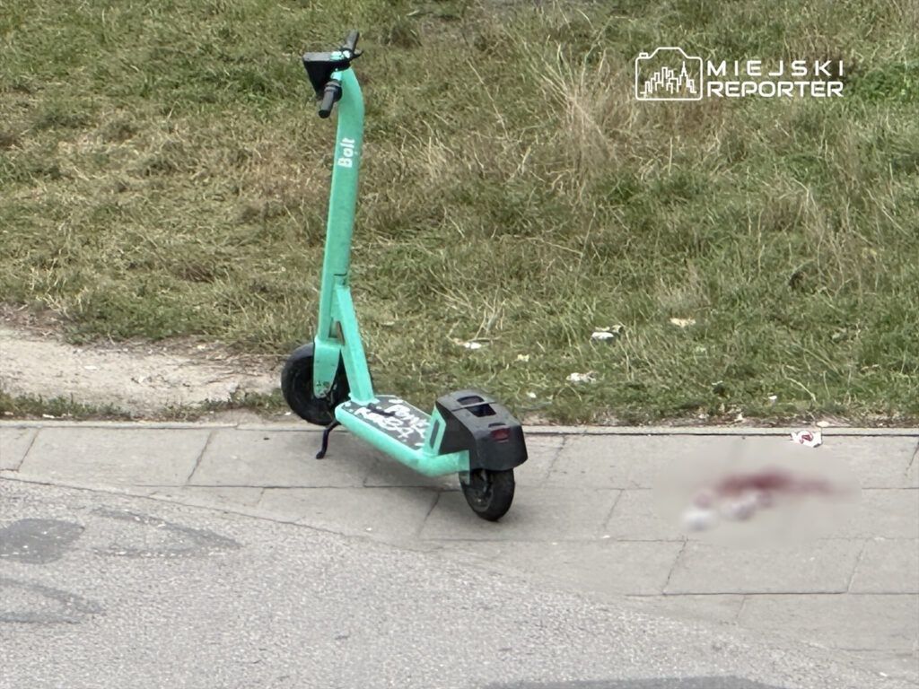 Zielona hulajnoga elektryczna stoi obok chodnika, obok niej widoczna plama na ziemi.