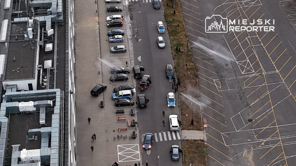 Grupa policjantów kontrolująca pojazdy na ulicy, w tle zaparkowane samochody i nowoczesny budynek.