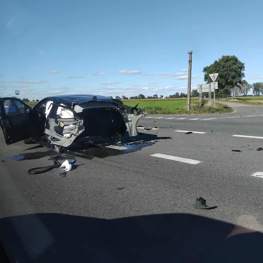 Zniszczony samochód leży na drodze, z otwartymi drzwiami, w otoczeniu pól i niebieskiego nieba. Wokół porozrzucane fragmenty pojazdu.