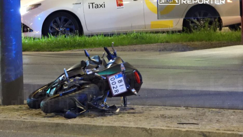 Zniszczony motocykl leży na chodniku obok słupa, a w tle widoczny jest taksówka z napisem "iTaxi".