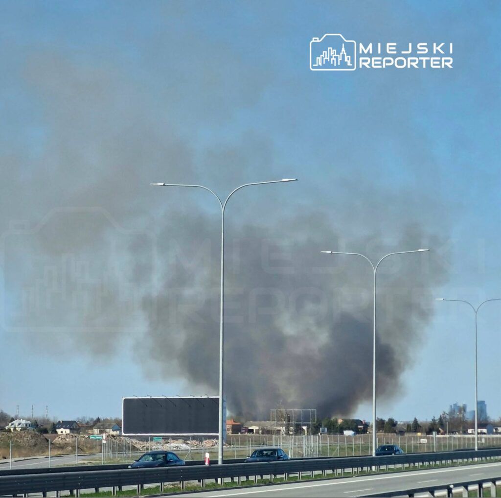 Chmura dymu unosząca się nad terenem zabudowanym, widoczna z drogi, obok stojące latarnie i billboard.