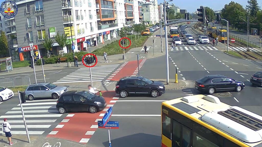 Grupa osób stojąca na chodniku przy przejściu dla pieszych, obok ruchliwej ulicy, w tle samochody i tramwaj.