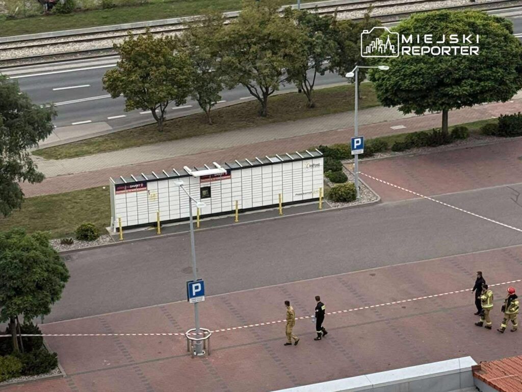 Grupa strażaków i policjantów spaceruje wzdłuż ogrodzonego terenu parkingu, w tle widoczne drzewa i torowisko.