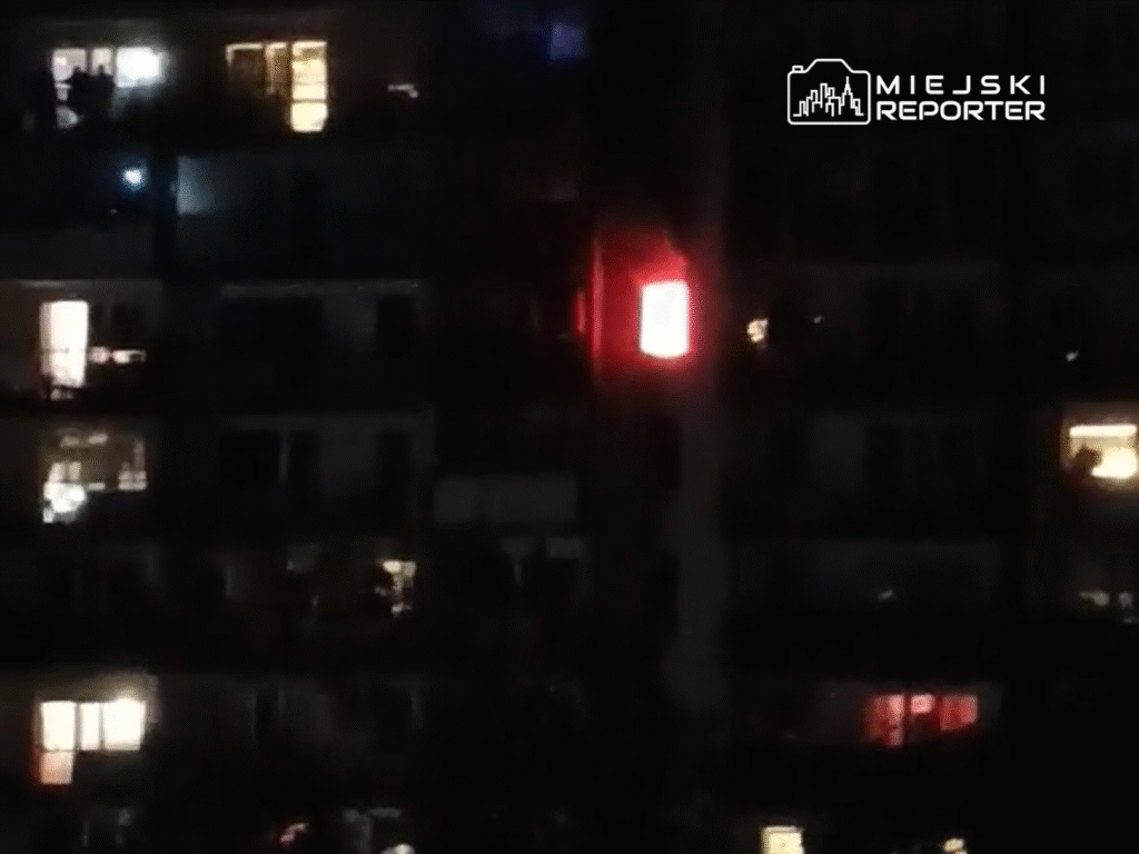 Oświetlony balkon na wysokim bloku mieszkalnym z widocznymi postaciami i czerwonym światłem w tle.