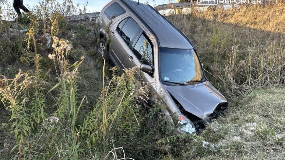 SUV w rowie, otoczony wysoką trawą i krzewami, obok strażacy przyglądają się sytuacji.