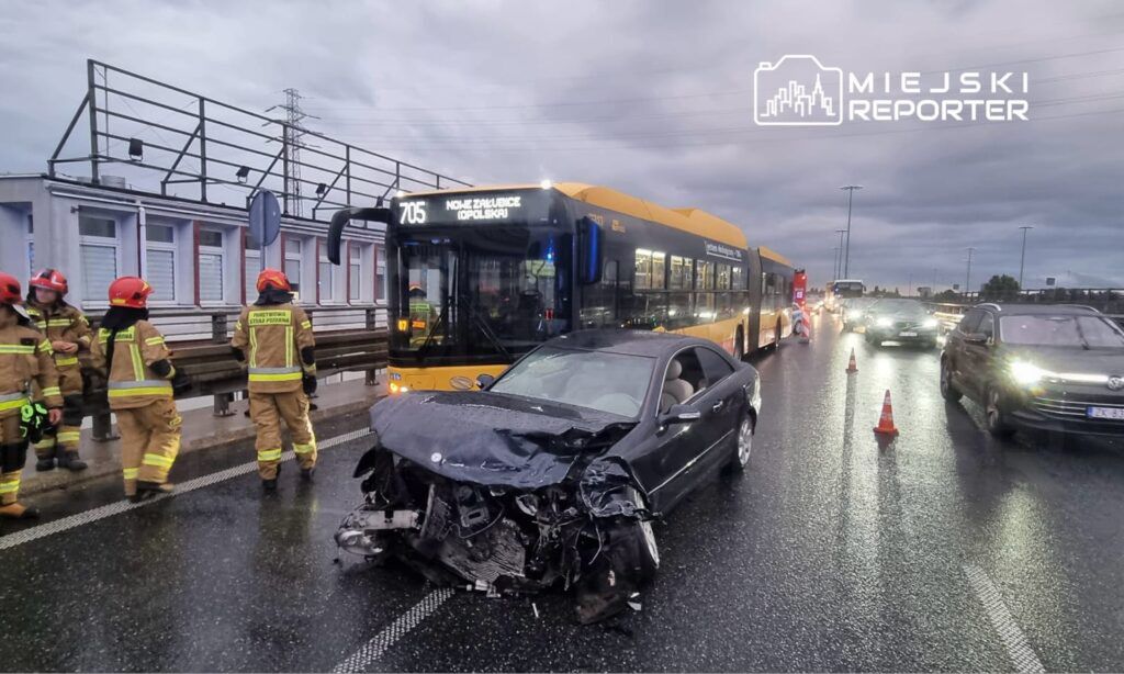Grupa strażaków w kombinezonach ratunkowych sprawdza uszkodzony samochód osobowy na jezdni obok autobusu komunikacji miejskiej.