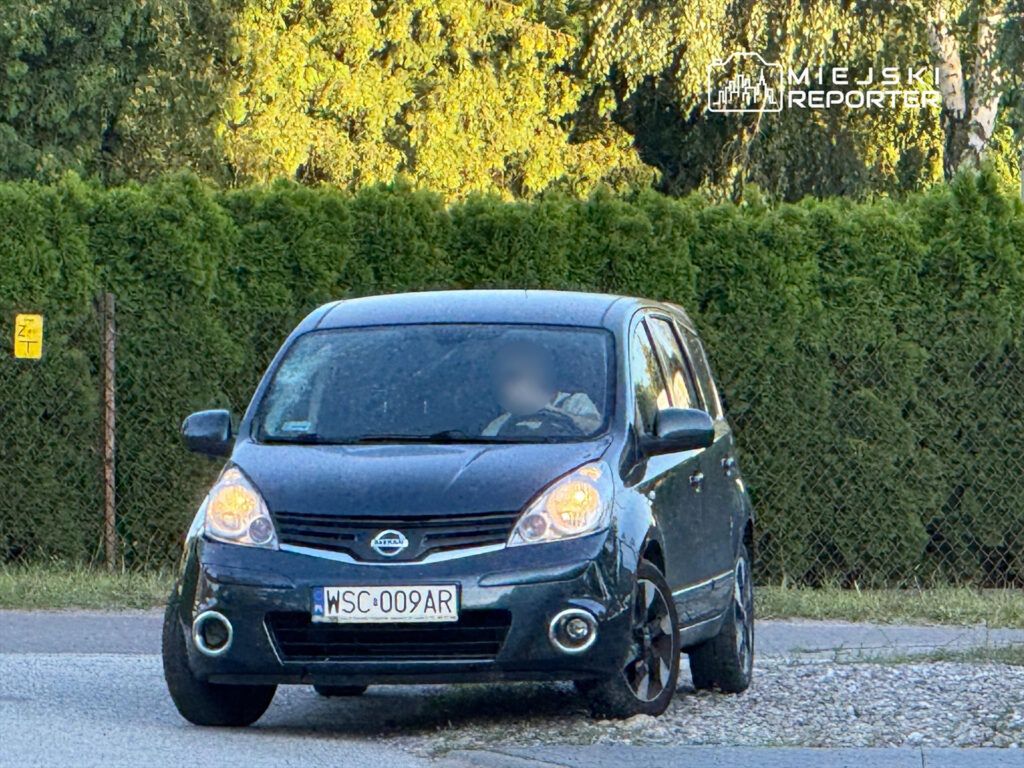 Czarny samochód osobowy zaparkowany na poboczu drogi, otoczony zielonymi żywopłotami.