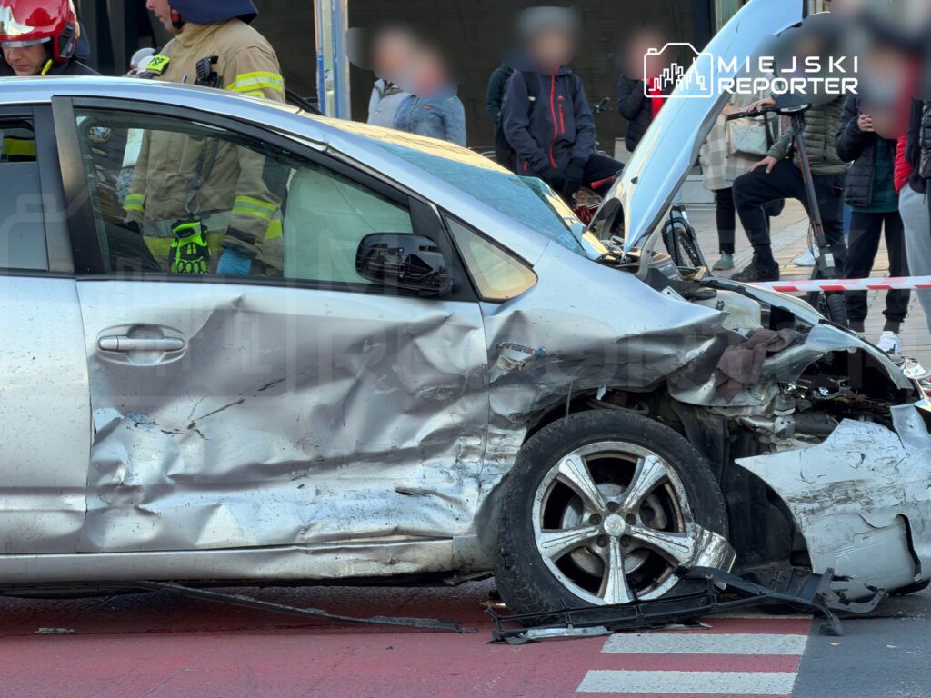 Zniszczony samochód osobowy z wgniecioną karoserią stoi na ulicy, obok grupy obserwatorów i strażaków.