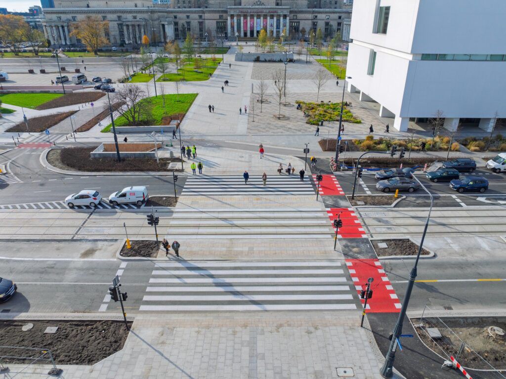 Grupa ludzi przechodzi przez pasy, obok nowoczesnego budynku i ruchliwej ulicy z samochodami.