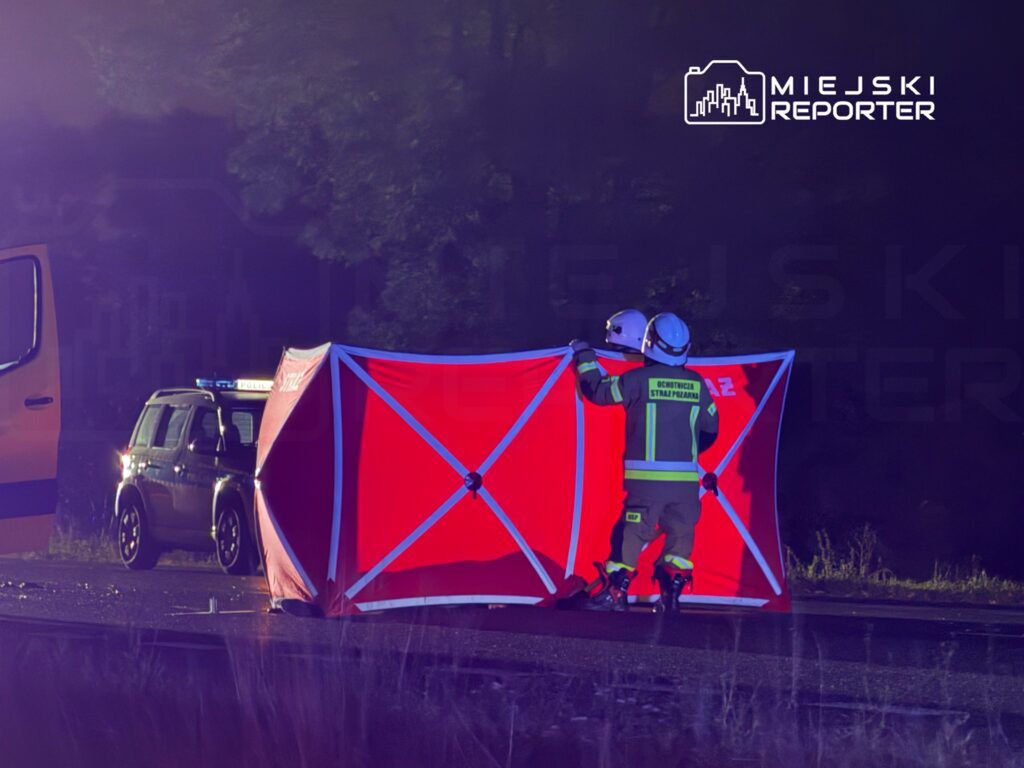Ratownicy w mundurach ustawią czerwony namiot na drodze, obok policyjnego samochodu, w ciemnym otoczeniu.